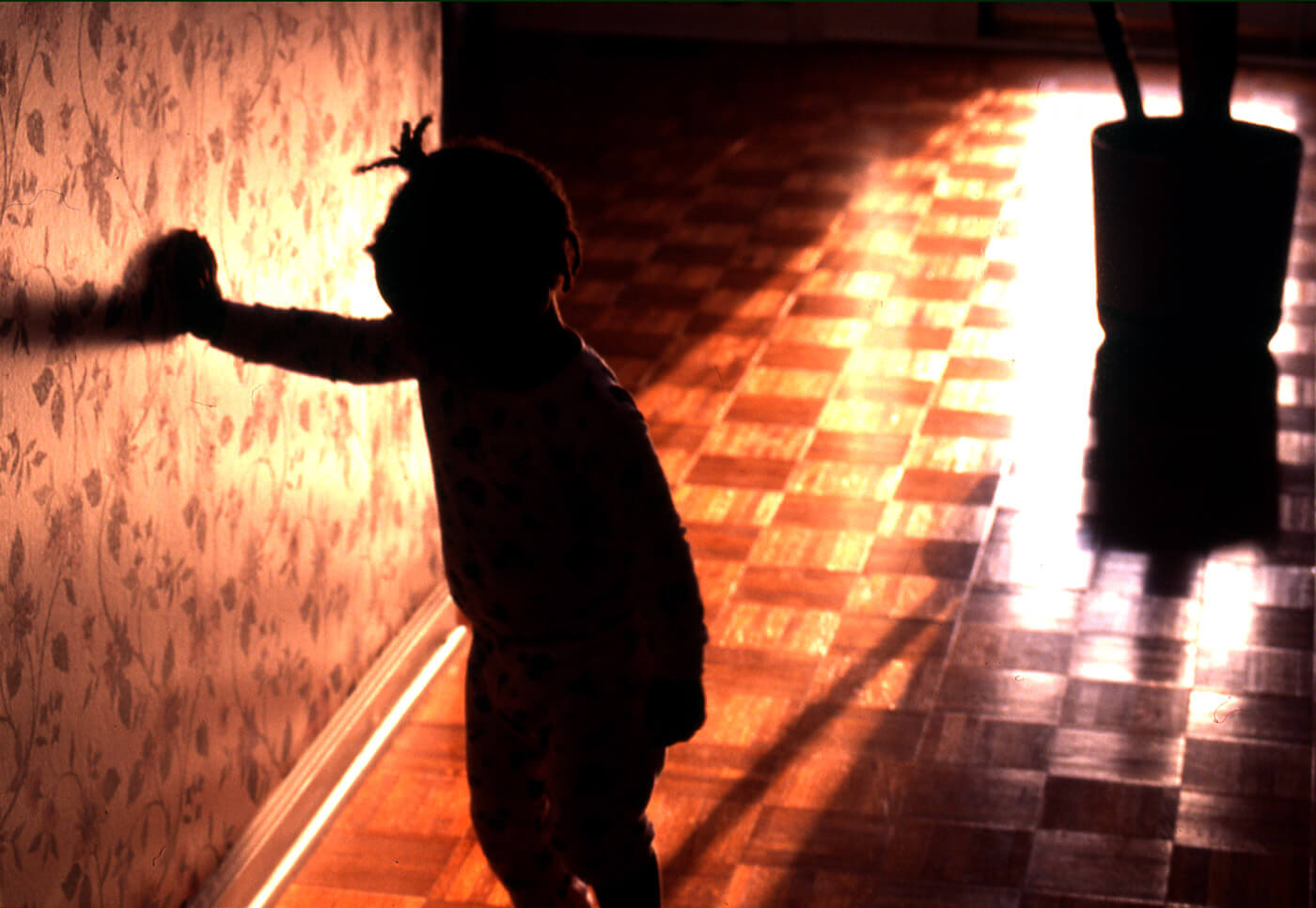 Silhouette of a child touching a patterned wall, framed by warm evening light across a wooden floor.