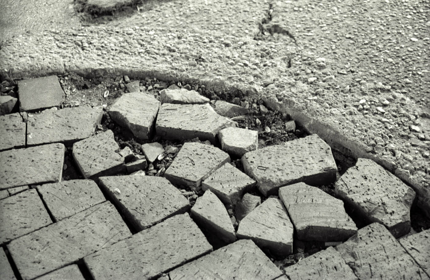 Close-up black and white photograph of broken sidewalk bricks and concrete, emphasizing urban texture.